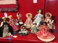 Group of 11 assorted collectible dolls displayed on a red surface, featuring Peggy Nisbet costume dolls, small Victorian style dolls, a Native American doll, and a Remembrance doll.