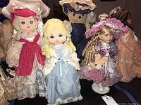 Group shot of four dolls: soft fabric dolls, porcelain doll with bonnet, and teddy bear in blue dress.