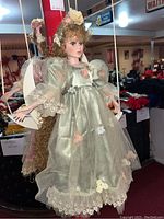 Close view of doll in pastel green dress with detailed lace and flower accents, curly hair with floral headpiece, hanging on crescent moon swing.