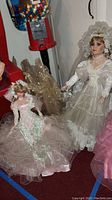 Two vintage porcelain dolls in detailed gowns; one standing in white lace bridal dress and veil, other seated in pink ruffled dress with pastel accents.