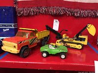 Photo showing all three Tonka toys on a red shelf: yellow dump truck, green car, and yellow excavator.