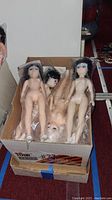 Photo of box containing multiple vintage plastic dolls new in packaging. Dolls have molded hair in black and white colors and are nude.