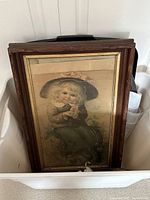 Photo showing framed antique artwork of a young child in distressed frame placed inside a white plastic bin.