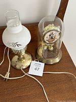 Antique brass electric lamp with frosted glass shade and anniversary clock with floral dial under glass dome on wooden surface.