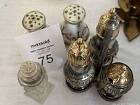 Set of six salt and pepper shakers including two Limoge ceramic, three metal and glass decorative, and one clear glass shaker with ornate top under moderate lighting.