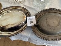 Two antique silver-plated serving dishes, one covered with a domed lid and floral edging, the other an open oval tray with rope-style border and handles, both tarnished.