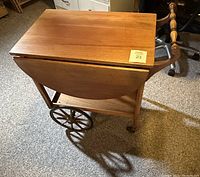 Side angled photo showing the tea cart with one drop leaf extended and one leaf down, two large rear wooden wheels and smaller front caster wheels visible, polished wood surface, and turned handle detail.