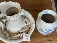 White ceramic wash basin with matching white pitcher inside, both featuring gold decorative accents, along with separate blue floral decorated pitcher.