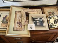 Photo showing a variety of framed small artworks including one with white tulips in a copper or metal frame, a framed ceramic bee trivet, other floral and botanical prints, and a wood rustic frame, all arranged on a wooden dresser.