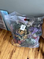 Clear plastic bag filled with assorted colorful artificial flowers on a wooden floor.