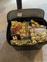 Open black storage bin filled with various Christmas decorations, primarily ornaments like Christmas balls in gold, red, and silver colors, bags of ribbons, and a gold candle holder visible inside.