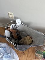 Oval metal coal scuttle filled with black metal scissors, wooden poker, and fire glove mitt, placed on wooden floor against wall.