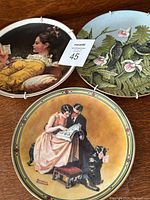 Three collector plates displayed on a wood table: Norman Rockwell 'A Couples Commitment', girl reading book plate, and birds plate.
