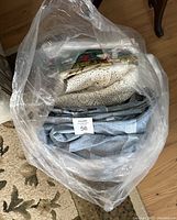 Top view of transparent plastic bag containing three folded textile items: cream crochet tablecloth, blue striped duvet cover, and patterned vinyl tablecloths.