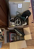 Top view photo of the lot items all together inside two boxes on a wood floor, showing a beige electric typewriter, black leather fanny pack, extension cords tangled, and several picture frames.