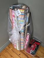 Photo of multiple rolls of assorted gift wrapping paper in plastic wrapping alongside a box of assorted ribbons.