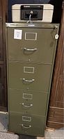 Front view of green 4-drawer legal size file cabinet with key attached and label holders visible on drawers, positioned between wooden furniture pieces in a basement.