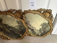 Pair of oval/scroll shaped plaster frames with gold finish and ornate detailing featuring shell motifs and floral carvings, holding artwork prints depicting a landscape with river, trees, and buildings.