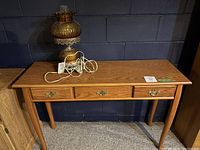 Full view of a rectangular faux wood sofa table with three drawers, brass handles, standing on four legs. A lamp (not included in sale) is placed on top.