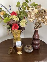Photo showing two floral arrangements; left arrangement is artificial flowers in yellow tin vase, right is dried flowers and pods in tall brown vase; amber candlestick holder and small decorative items placed in front.