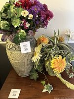 Photo showing a large, heavy ceramic vase with textured decorative patterns filled with artificial flowers including purple and white flowers and green leaves. Beside it is an additional floral arrangement of yellow flowers and greenery on a wooden surface.