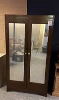 Front view of vintage wood armoire with two full-length mirrored doors, brass/gold knobs, reflecting basement environment.