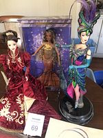 Three Barbie dolls displayed: Diva in red/gold gown, Holiday Ball in gold dress (boxed), Tango Barbie in colorful Bob Mackie outfit with feathers on a black stand