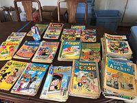 Wide top-down photo of multiple stacks of vintage comic books including Little Dot, Betty & Veronica, Richie Rich, Little Archie, Archie, Hot Stuff, Casper, Sabrina, Blondie, The Super Friends, Dennis the Menace, and more, plus a Beatles nesting doll in the center.