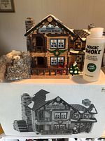 Front angle view of the Department 56 Smokey Mountain Retreat house with wood pellets and Magic Smoke pine scent bottle displayed beside it. Black and white box image shown below the house.