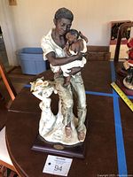 Full view of the porcelain figurine showing the man sitting with the child wrapped in cloth, mounted on wooden base.