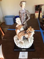 Full view of handcrafted detailed porcelain figurine showing lady in period dress with two Borzoi dogs at her feet, mounted on black base.