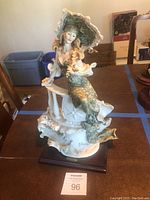 Full side view of figurine seated holding parasol on wooden base with MaxSold tag 96.