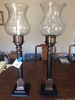 Pair of two hurricane lamps side by side on wood table, showing full height and metal and glass construction