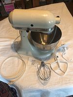 White vintage KitchenAid stand mixer with stainless steel bowl, clear splash guard, and three attachments (whisk, dough hook, flat beater) laid out on a white table.