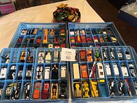 Photo showing two full blue plastic carrying cases containing multiple individual Matchbox cars and vehicles organized in compartments, plus a separate round container with miscellaneous loose cars and vehicles.