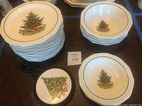 Stack of 16 dinner plates with Christmas tree design, 4 serving bowls, 2 soup bowls, and black metal trivet with a decorated Christmas tree image