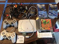Overview photo showing PS One console, two controllers, hookup cables, power adapter, extension cable adapters, and game discs including Frogger 2.