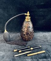 Photo of large lustre-ware perfume atomizer with black bulb pump and tassel next to four vintage pendant pens and pencils in varying designs and gold accents.