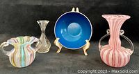 Four small decorative glass pieces on black background: two pink and white ribbon swirl vases, one clear etched bird vase, one blue bowl on stand
