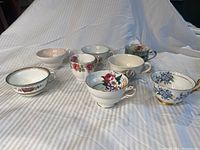 Photo showing all eight assorted teacups arranged in two rows on striped cloth background under natural lighting, visible varied floral patterns and gold trim.