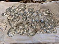 Various cut clear glass and crystal chandelier drops and prisms arranged on a white cloth, showing teardrop, oval, octagonal, and irregular shapes, some with metal hooks.