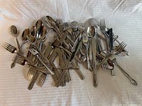 View of assorted forks, knives, and spoons spread out on a white cloth with mix of simple and decorated metal handles.