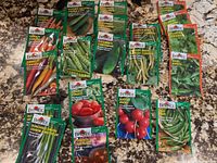 Photo of assorted seed packets laid out on countertop including carrots, onions, peas, radish, herbs, tomato, pumpkin, cucumber, zucchini, and beans from Burpee brand.