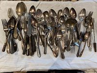 Metal cutlery assortment including forks, spoons, some serving utensils laid on cloth. Items appear tarnished and mixed styles.