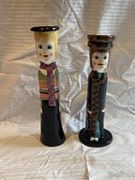 Front view of two ceramic vases depicting stylized human figures; one with leopard coat, one with scarf and hat.