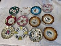 Top-down view of all 11 decorative saucers with varied colorful floral and patterned designs, arranged on a white cloth background.