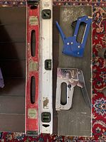 Two bubble levels and two staple guns laid out on surface with colorful rug beneath.