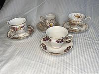 Four bone china teacups with matching saucers, each with floral patterns and gold trim, displayed on a white striped fabric.