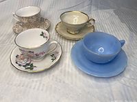 Four teacups with matching saucers set on a gray fabric background showing different styles and colors.