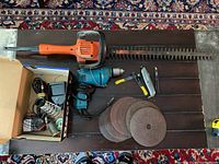 Black & Decker hedge trimmer with orange handle, electric Makita drill with cord, staple gun with yellow and black handle, six sanding discs on wooden surface